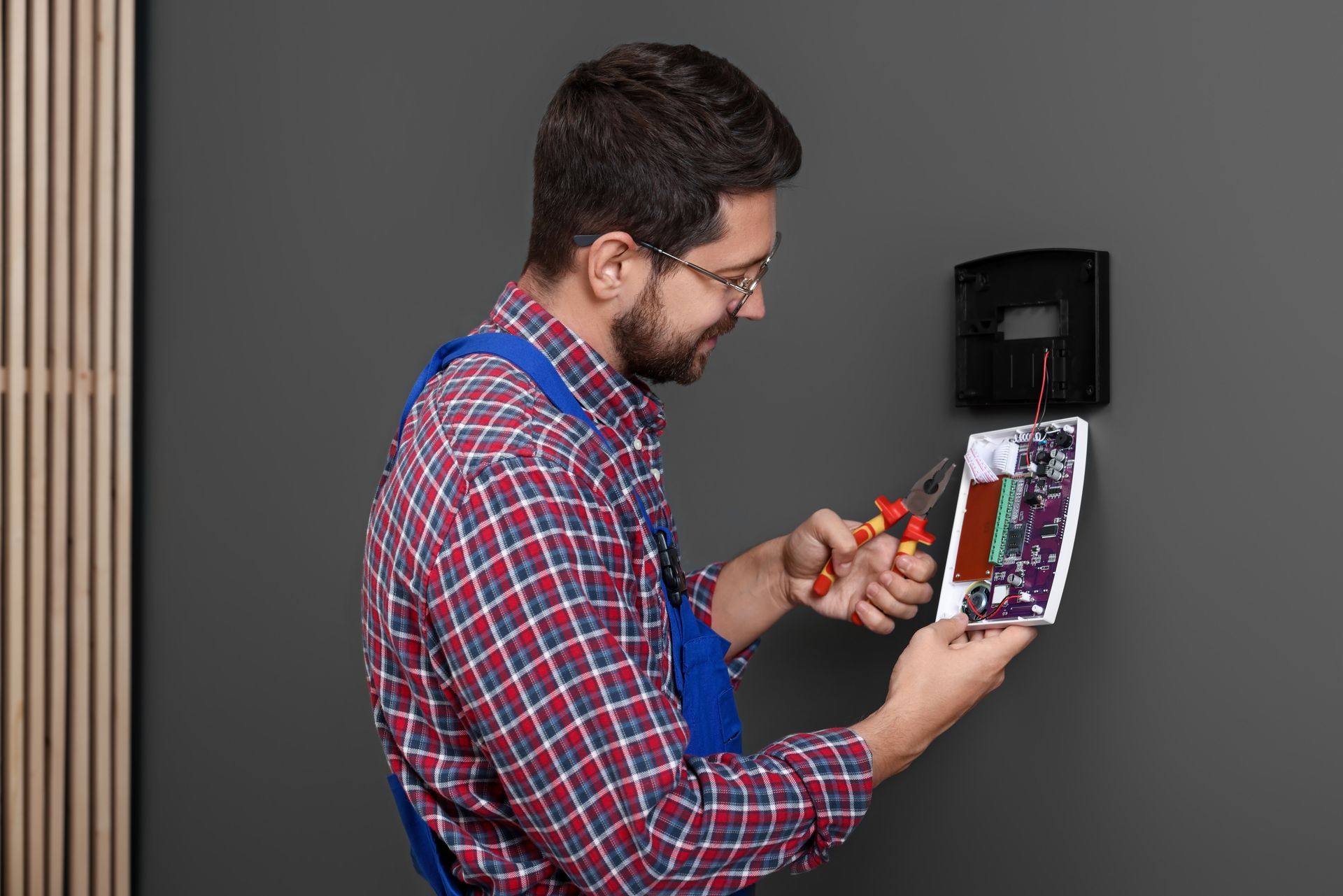 Man in overalls and glasses using pliers to work on a circuit board on a wall.