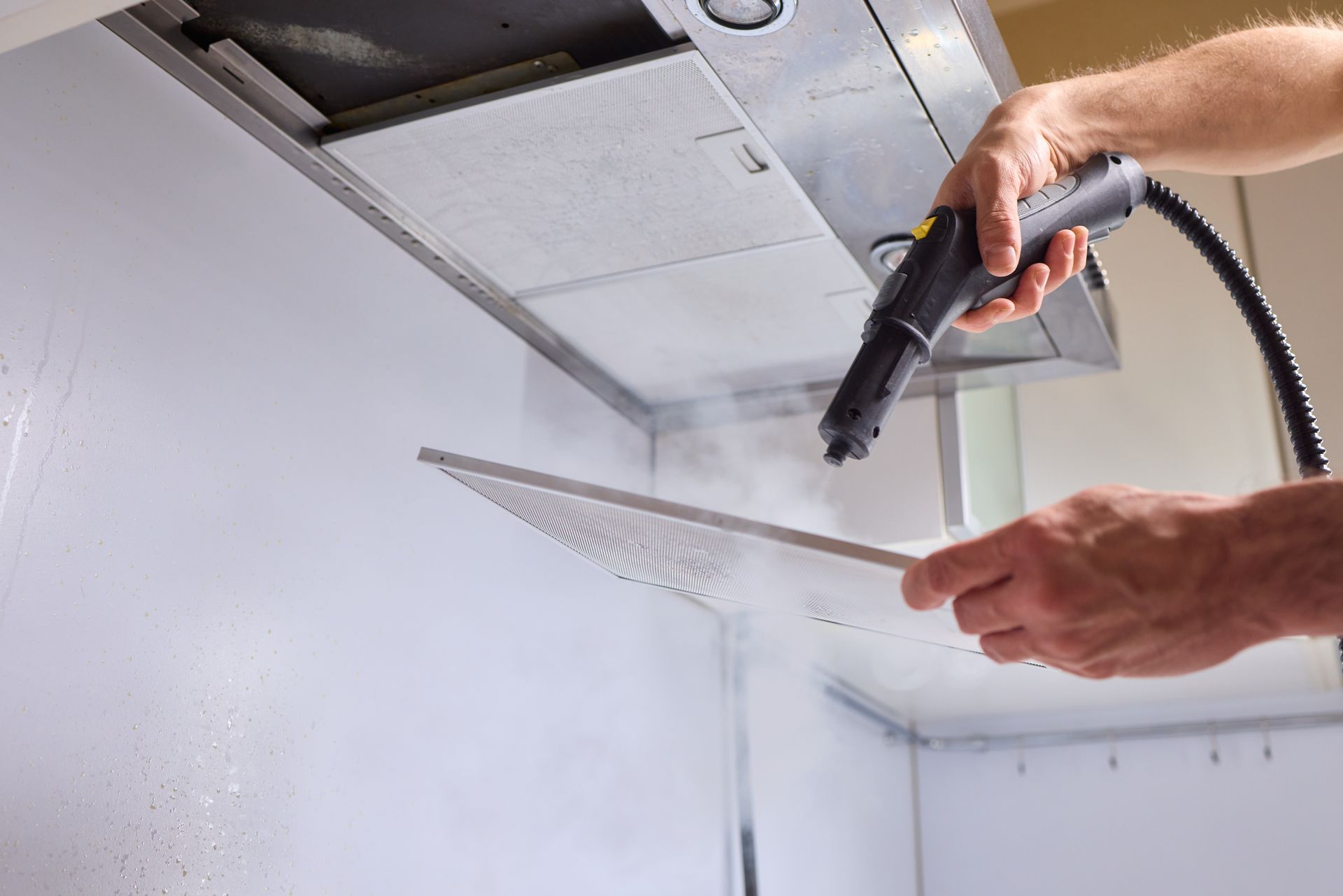 Person cleaning a metal filter with a steam cleaner under a kitchen hood.