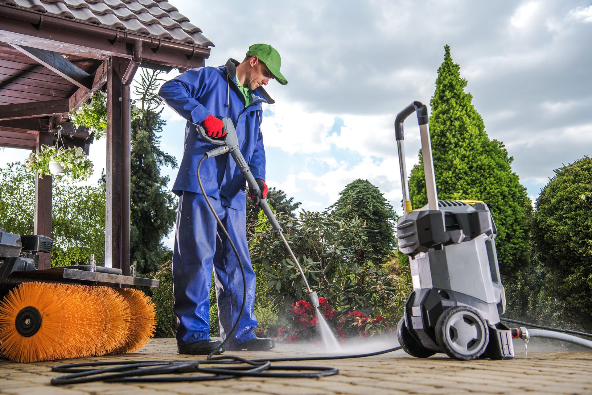 Person pressure washing a patio, using a power washer with a hose and wearing protective blue coveralls and a green hat.