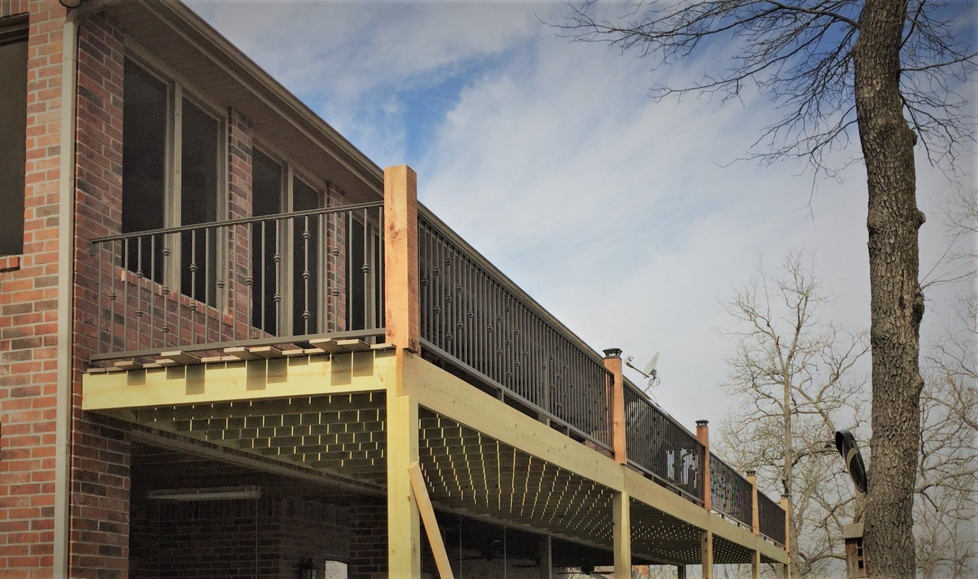 Brick house with a raised wooden deck and stairs beside leafless trees under a cloudy sky