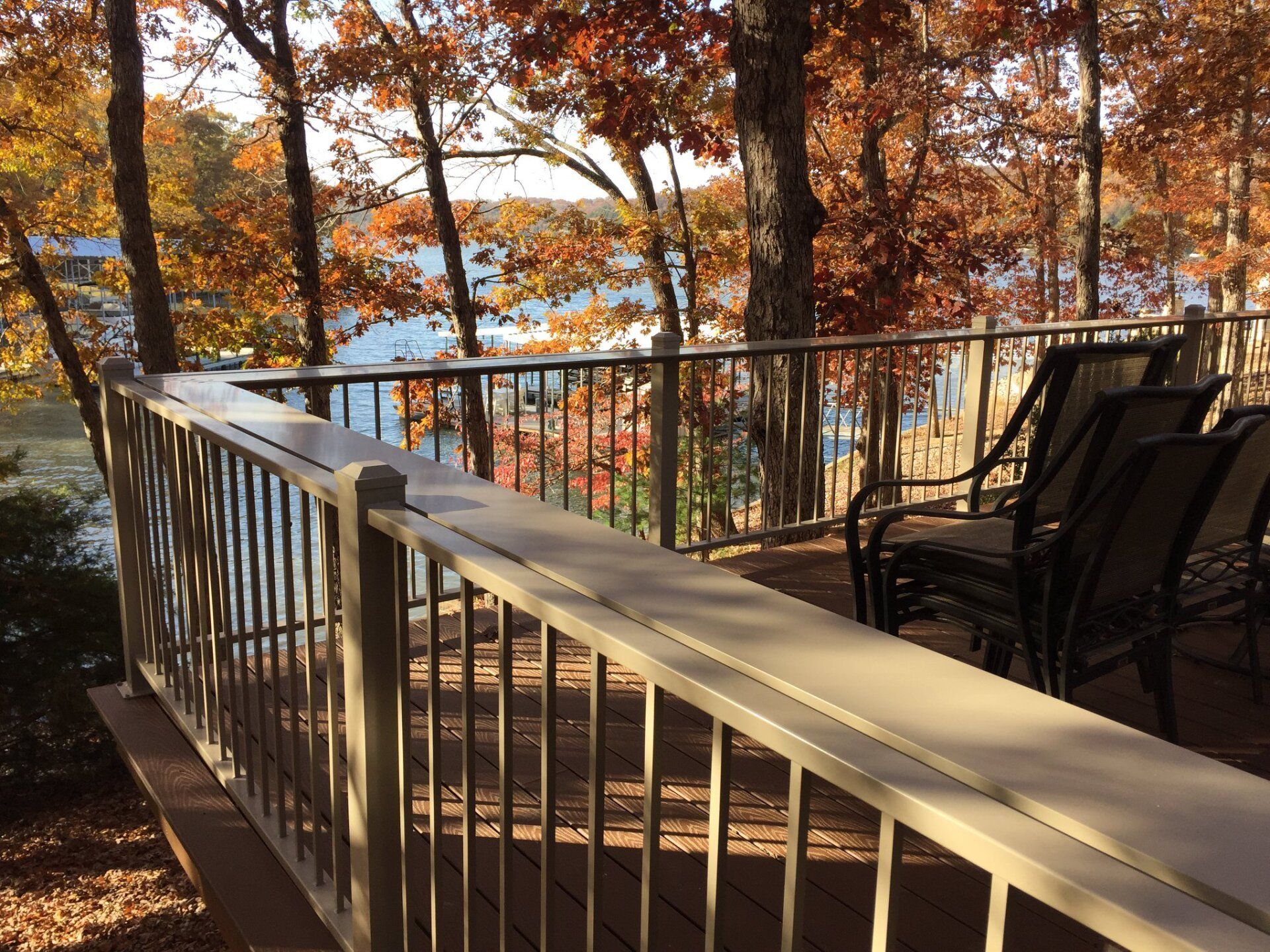 Sunlit deck with white railing, black chairs, and autumn trees overlooking water