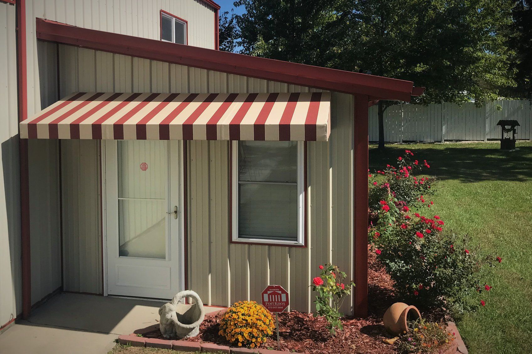 Small building entrance with red-and-white awning, white door, window, and flower bed in front