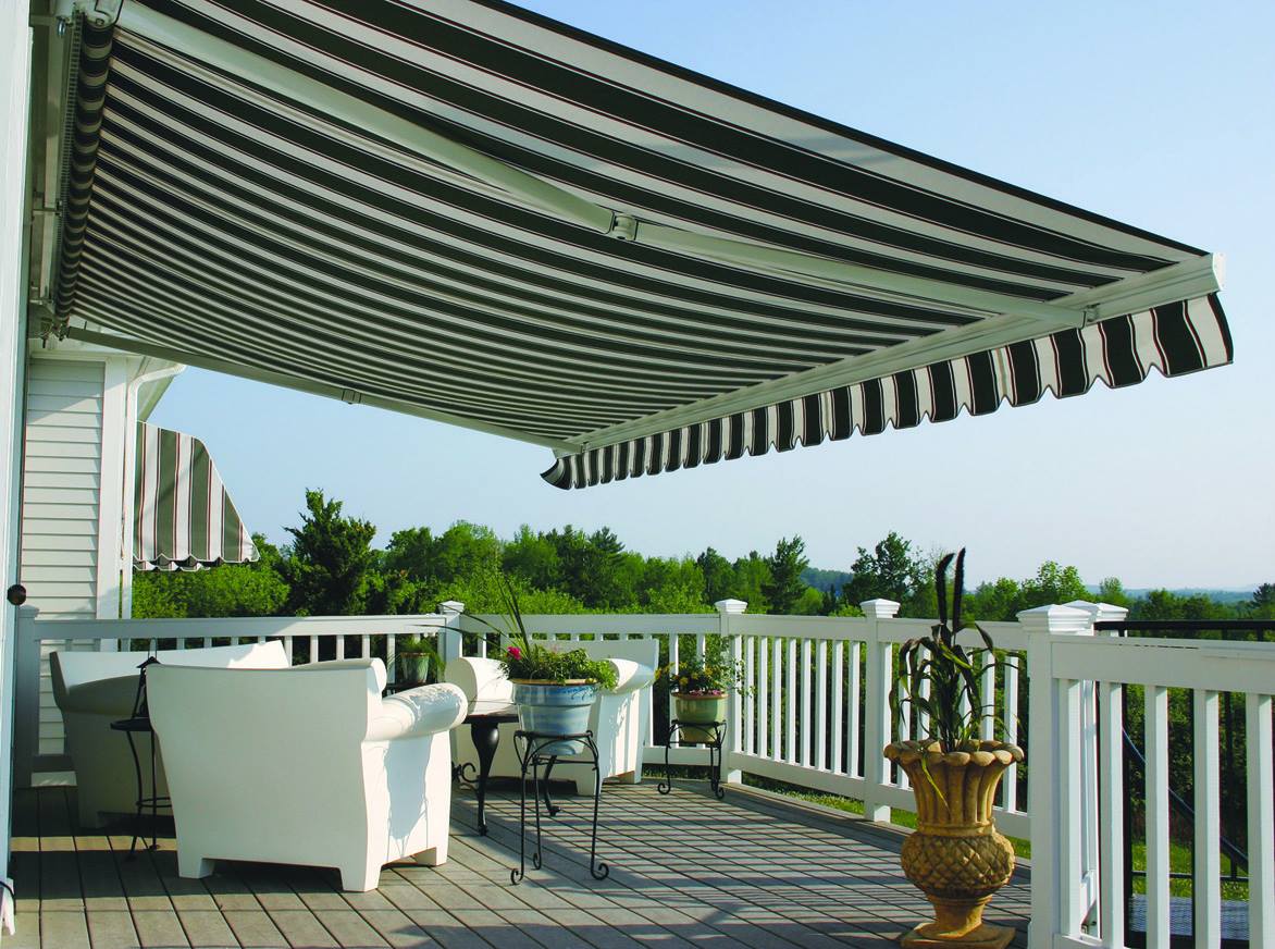 Patio with striped awning, white chairs, potted plants, and a railing overlooking greenery and water.