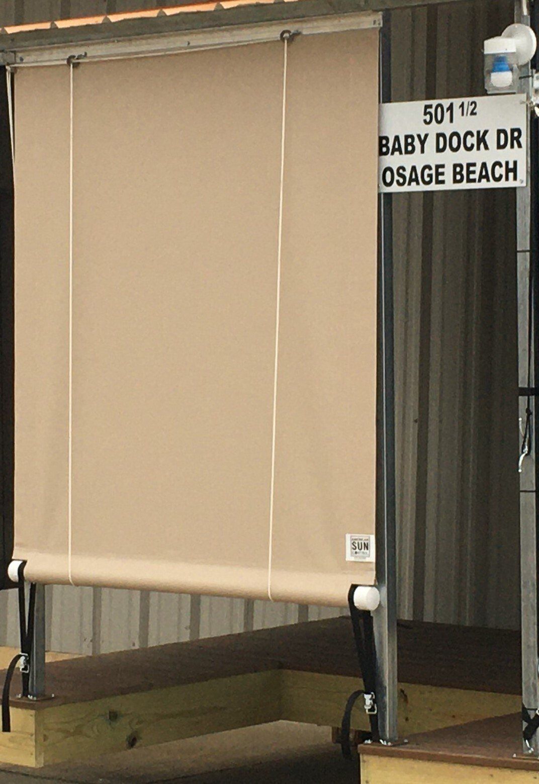 Closed beige roll-down shade beside a black sign reading “BAY DOCK DR, OSAGE BEACH.”
