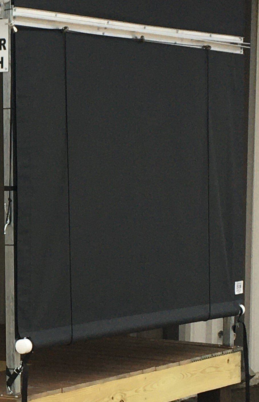 Black roller blind over a wooden platform frame in a room, with a white ball weight on the left cord.