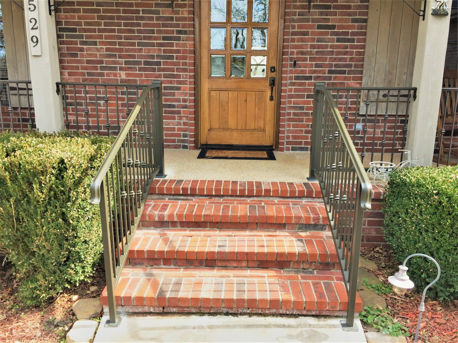 Brick front porch with wooden door, brick steps, and metal railings bordered by shrubs.
