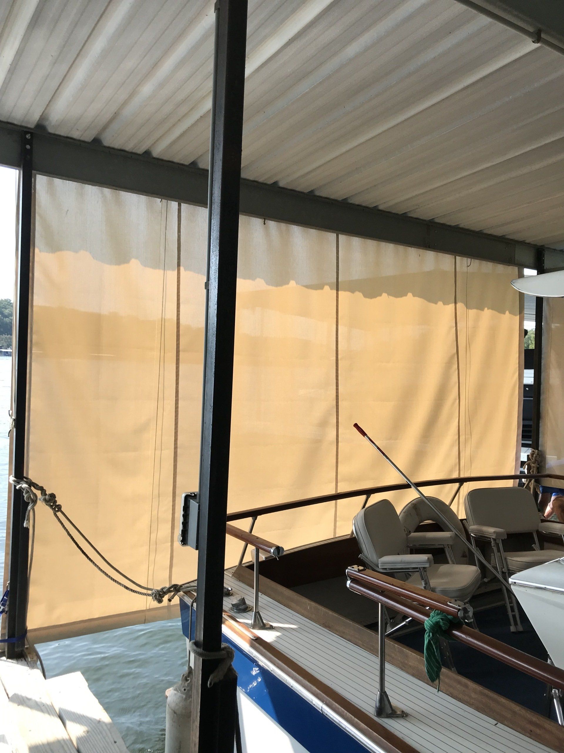 Covered dock with black posts, chain railings, and tan window shades beside the water.