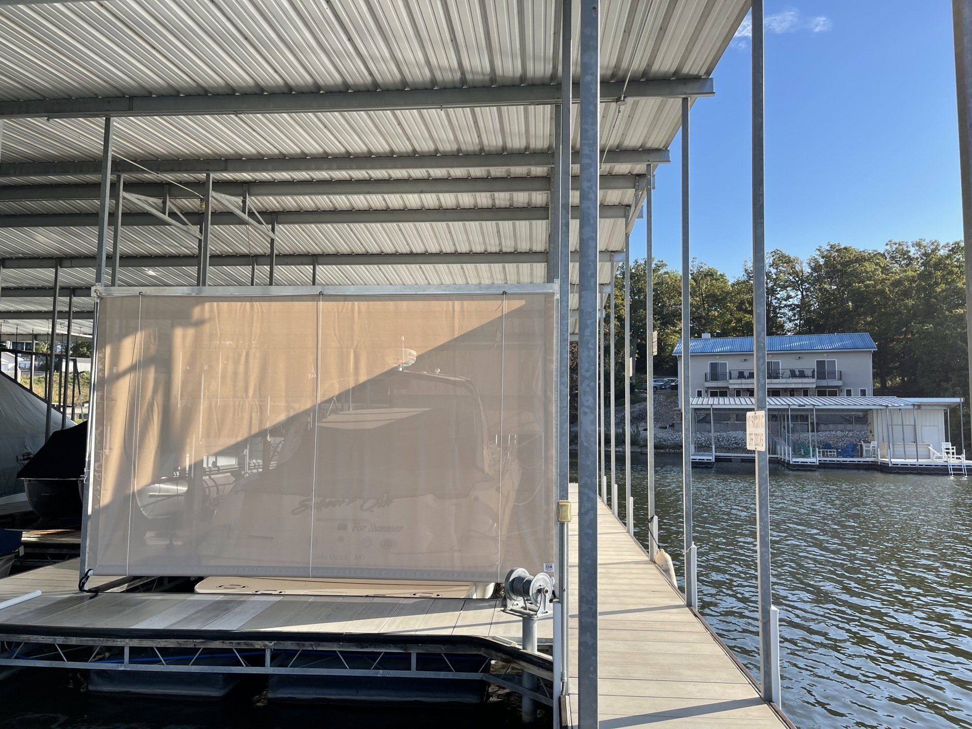 Covered dock with a tan screen, metal roof, and calm lake beside a building in the background