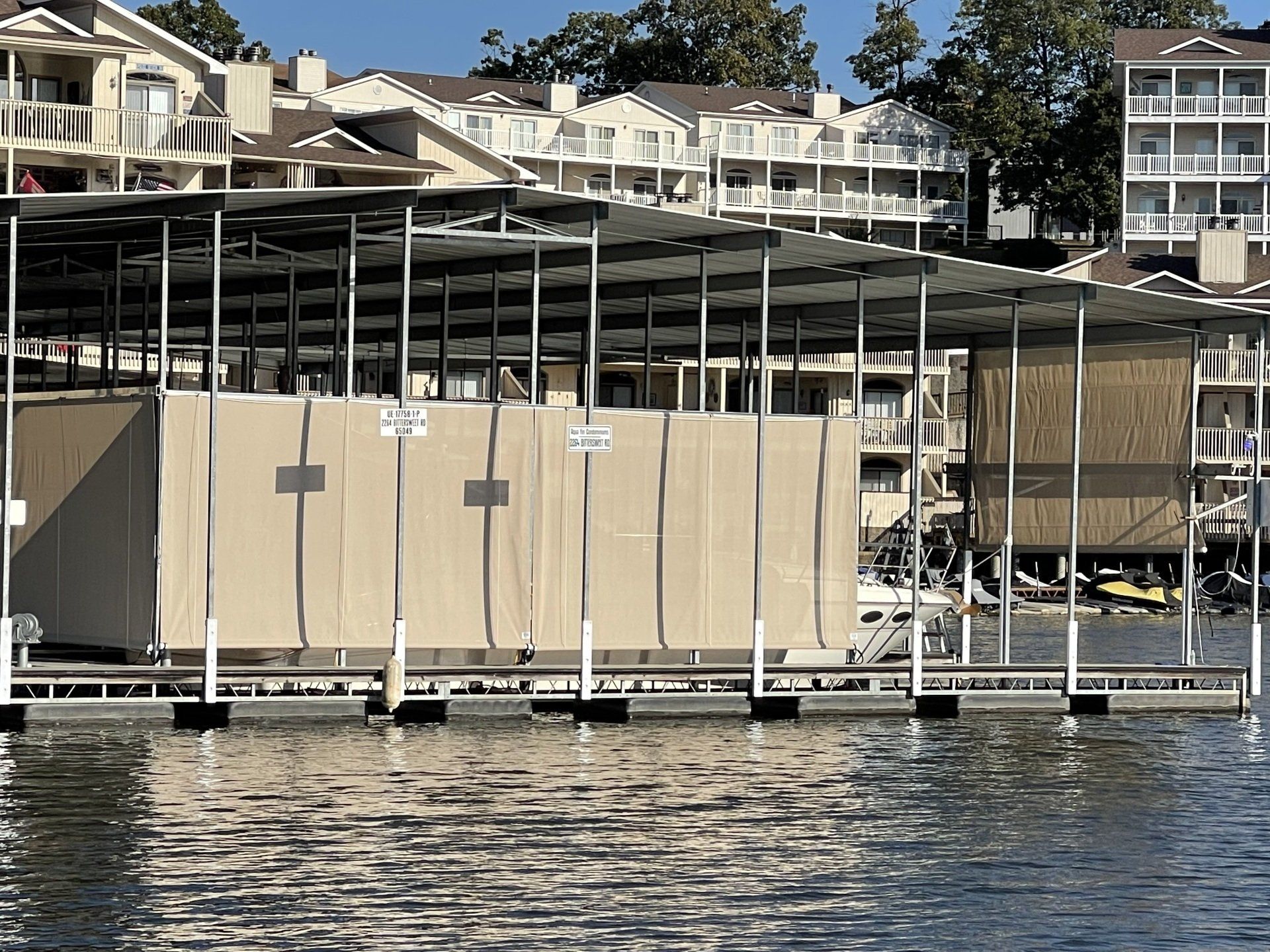 Covered boat slips at a marina with beige curtains, water in the foreground, and houses on a hillside behind.