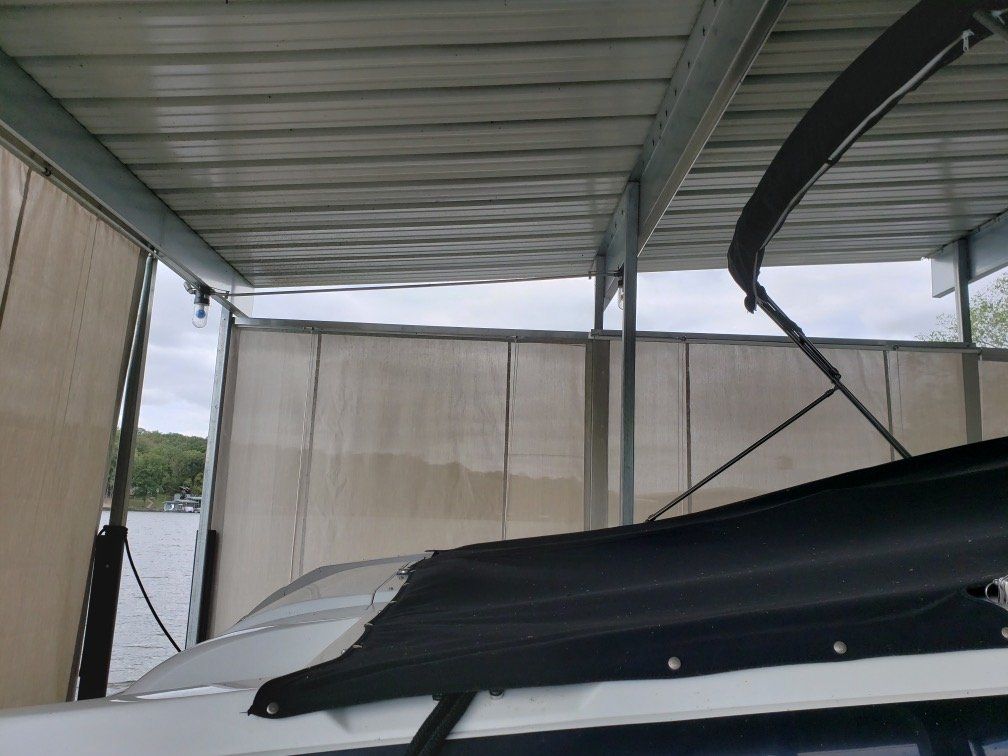 Covered boat docked under a metal roof by tan privacy screens, with water visible beyond.