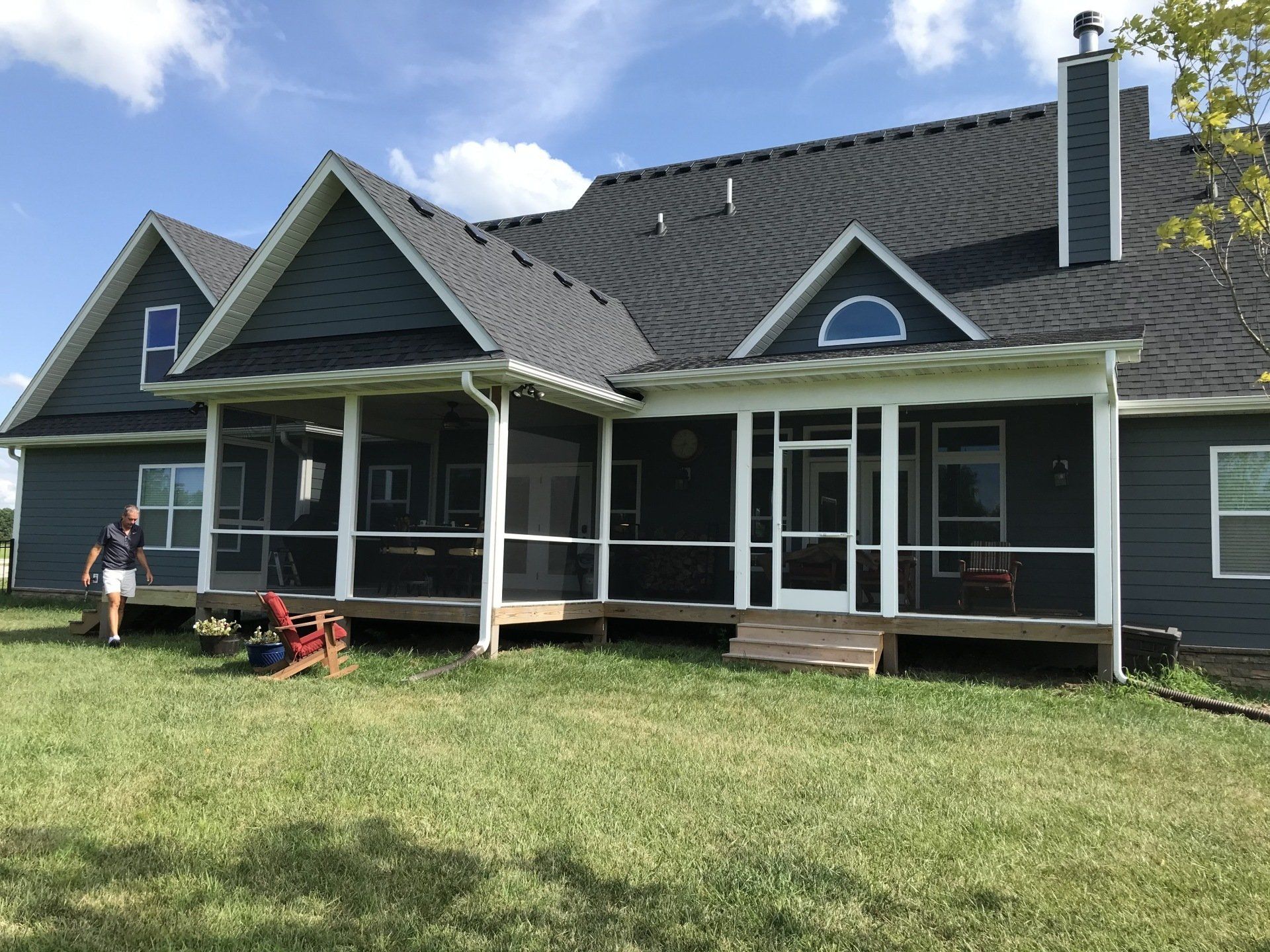 House with dark roof, screened porch, and two people in lawn chairs on a grassy yard under a blue sky