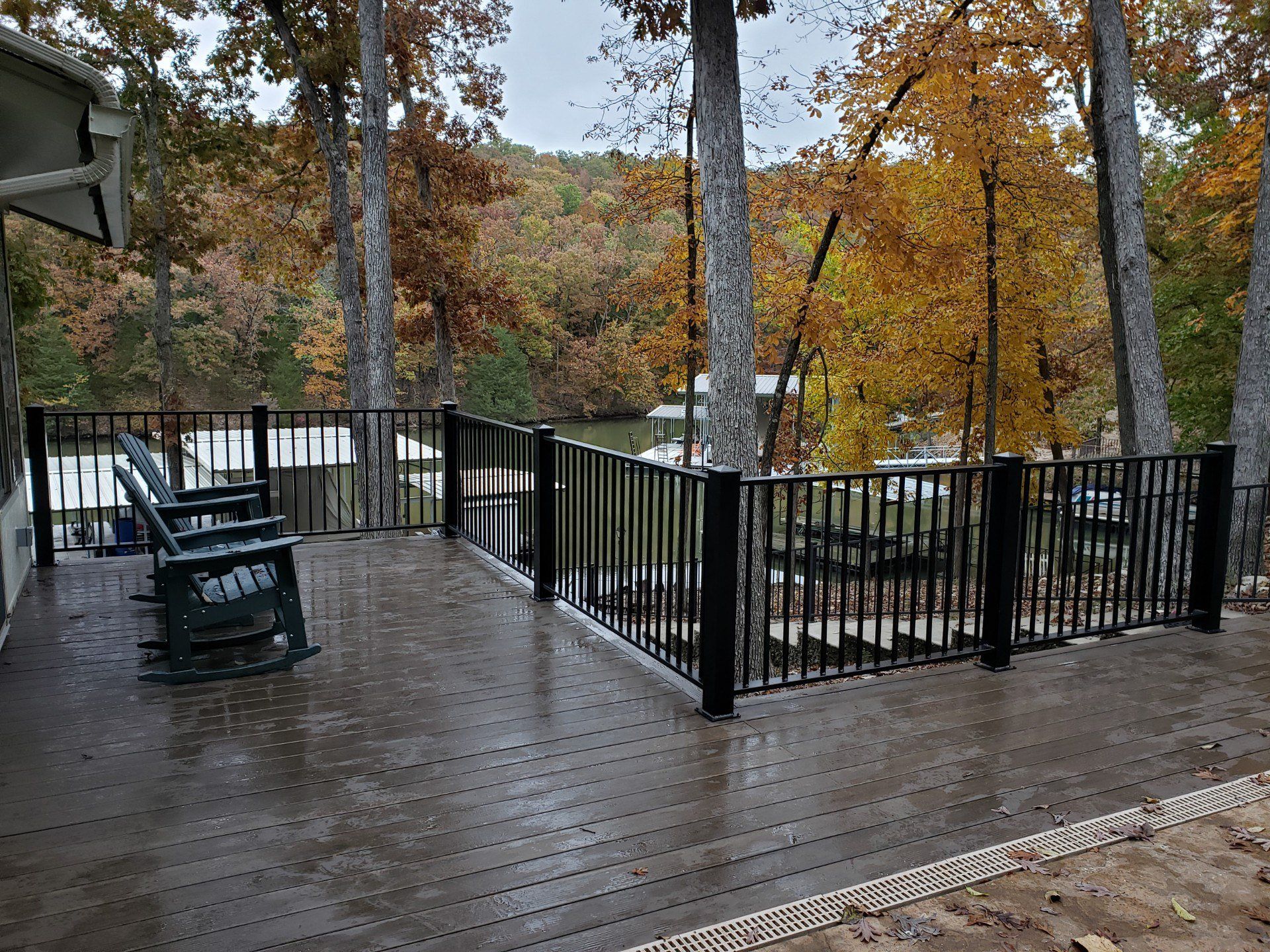 Rainy wooden deck with black railings, chairs, and a fire pit overlooking autumn trees and a lake