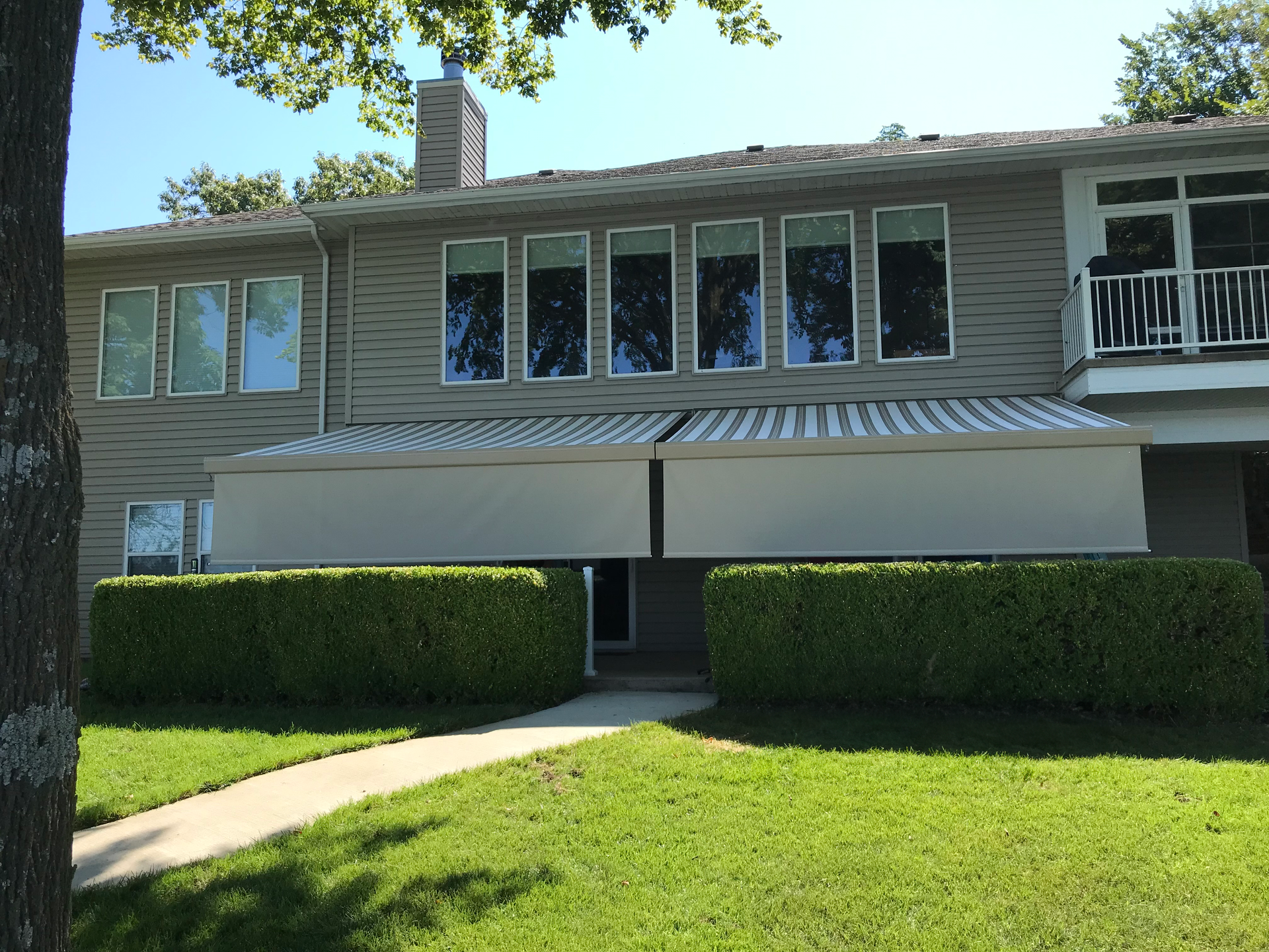 Two-story house with striped awnings, trimmed hedges, and a green lawn on a sunny day