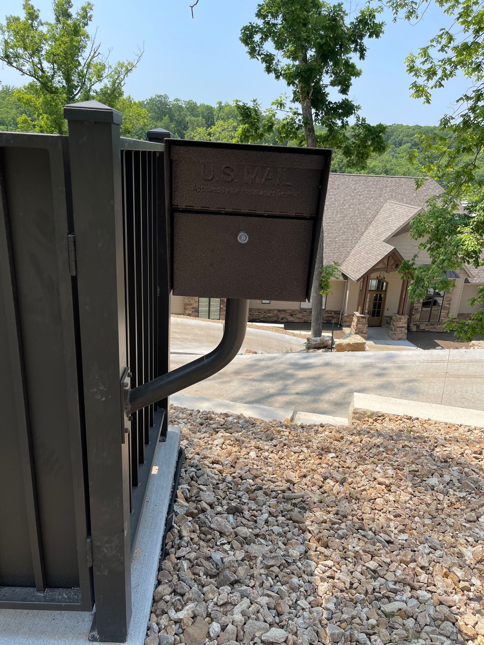 Black fence with a mounted mailbox beside a gravel driveway and house in the background.
