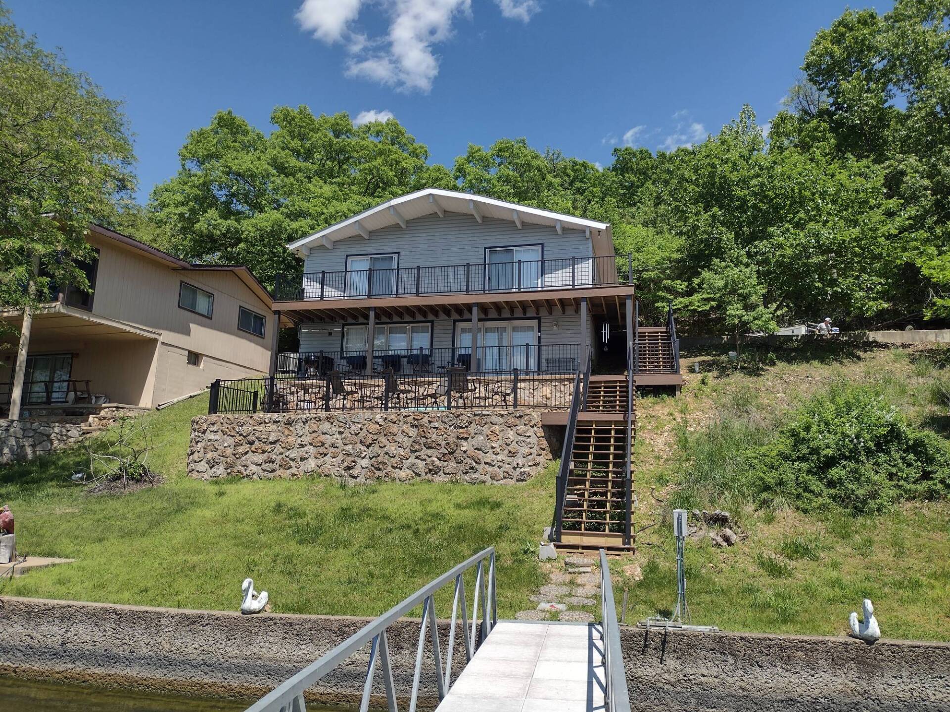 Two-story lakeside house with stone retaining wall, wooden deck, and steps down a grassy slope under a blue sky