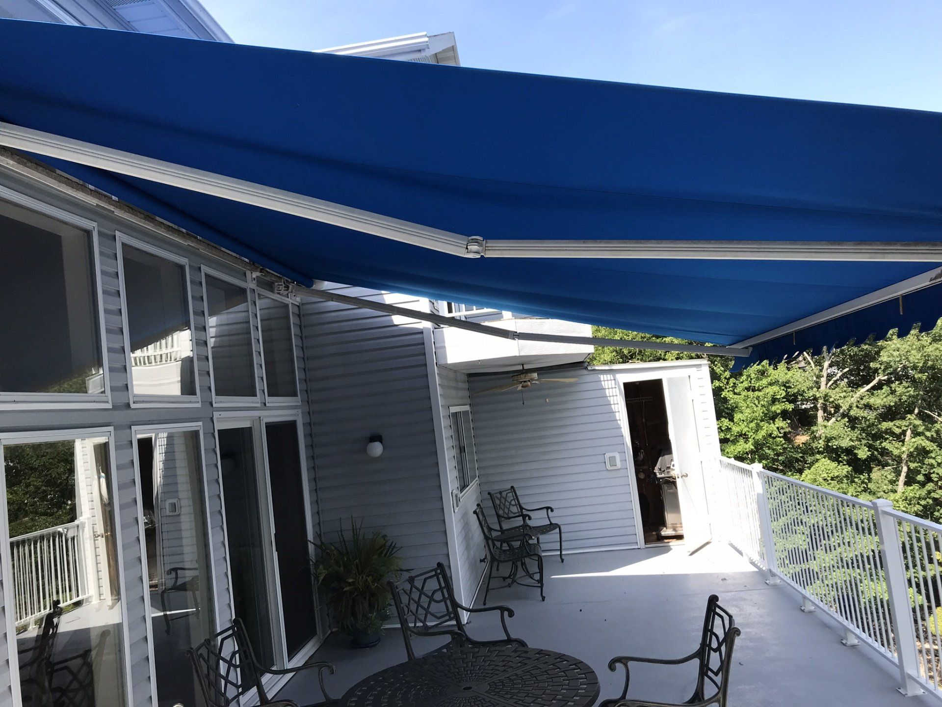 Covered patio with blue awnings, white railing, and outdoor chairs beside a house.