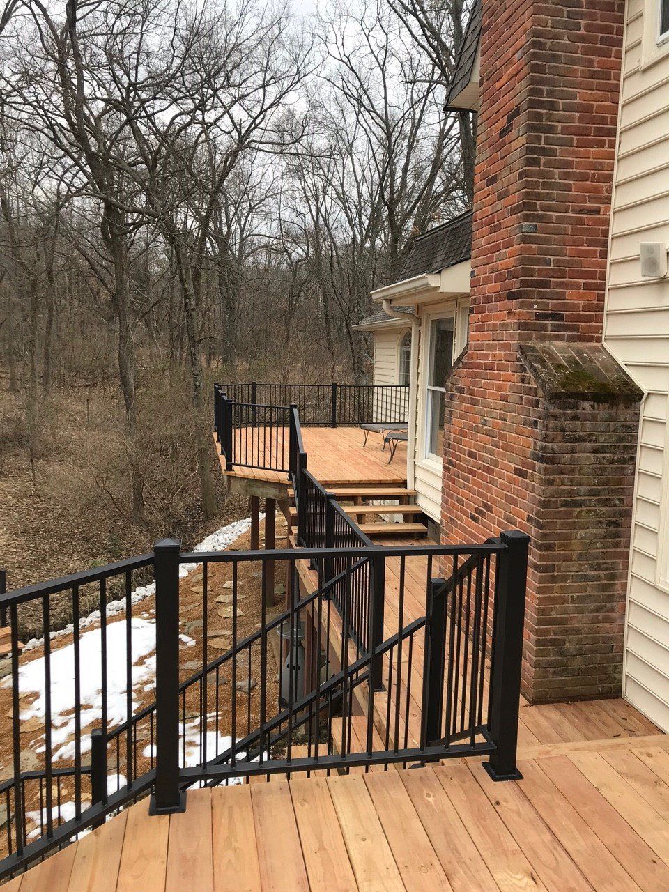 Wooden deck and black railing beside a brick house, overlooking a wooded yard with patches of snow