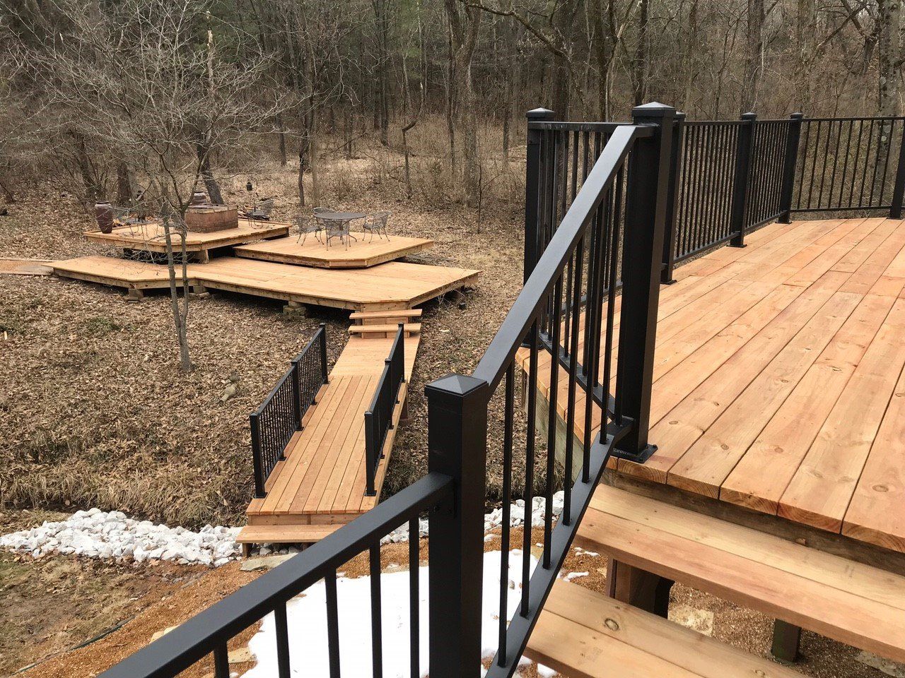 Wooden deck with black railing overlooking a narrow wooden footbridge in a wooded area