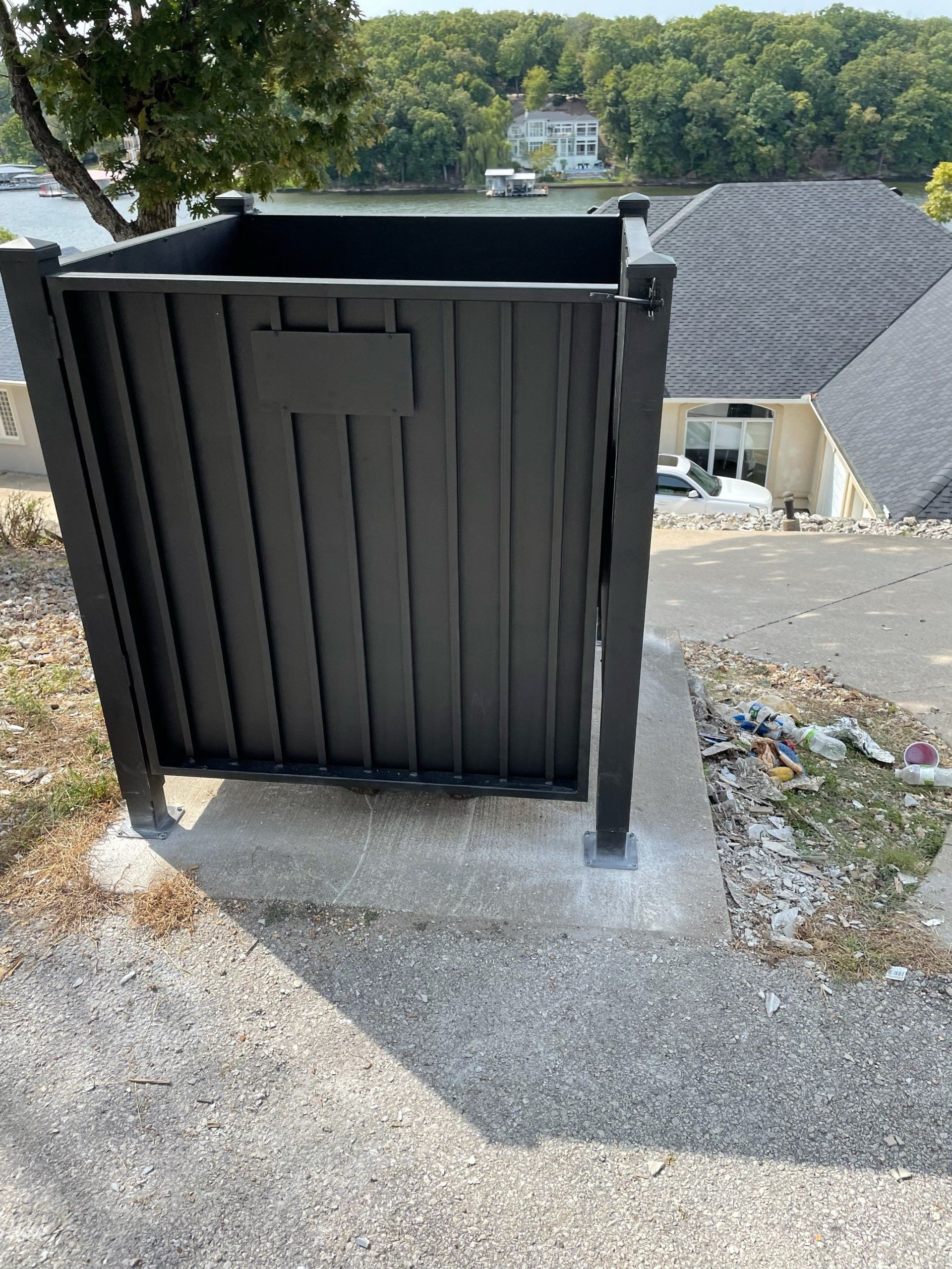 Black utility enclosure on a concrete pad beside a house, overlooking a wooded hillside.