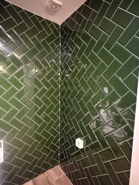 A corner shower wall featuring glossy, dark green subway tiles arranged in a herringbone pattern with white grout.