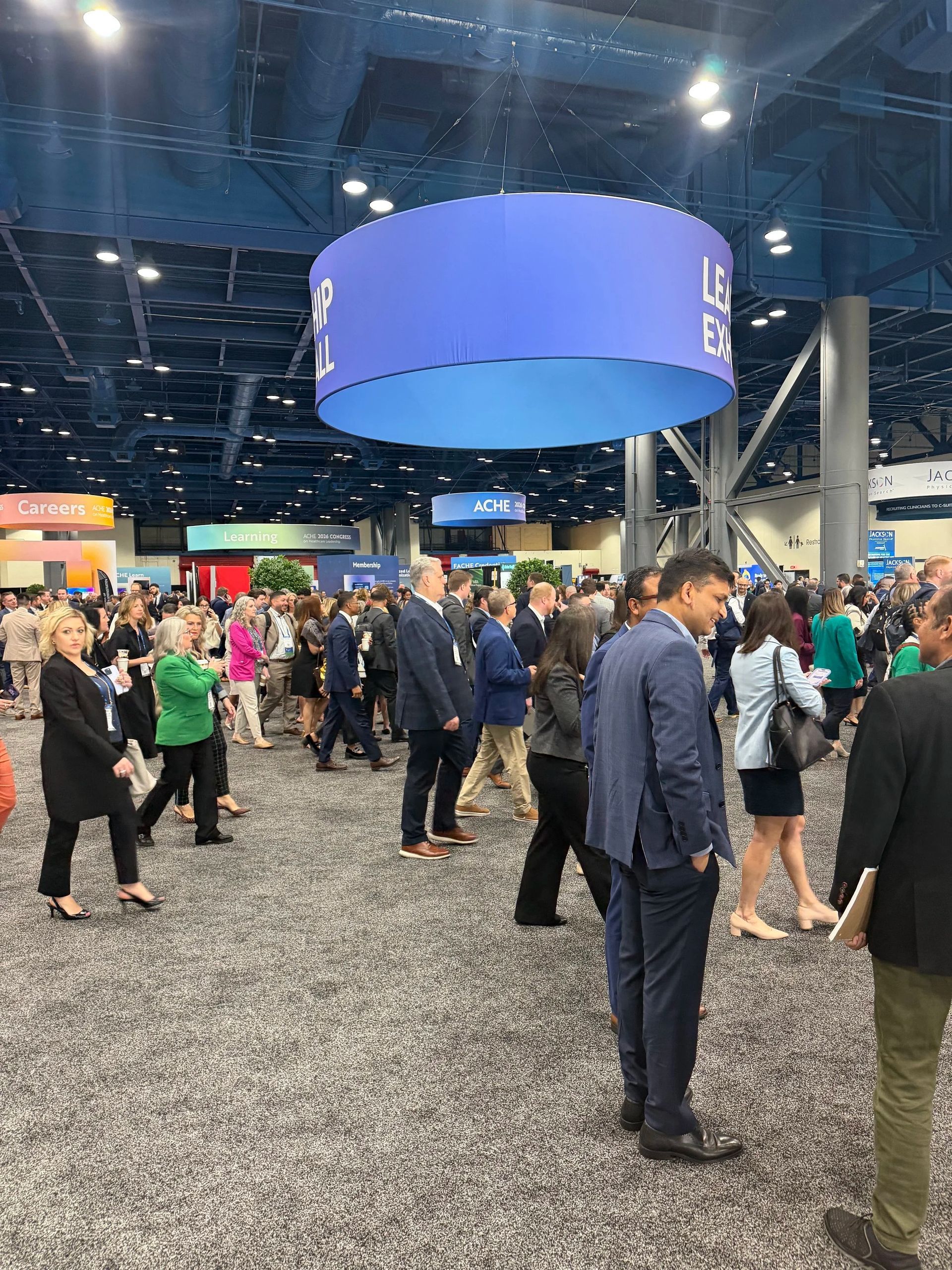 Crowded convention hall with people standing beneath a large blue hanging sign.