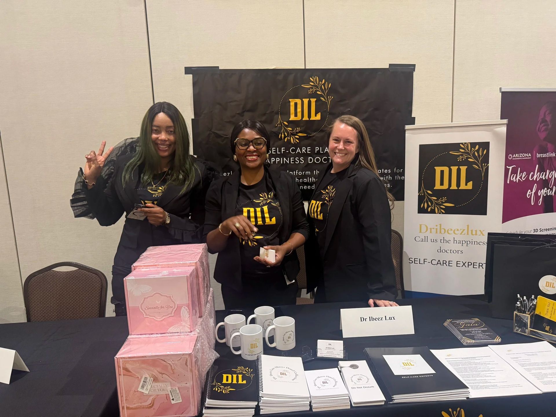 Three people at a booth with Delta Iota Lambda banners, tables of pamphlets, and pink gift boxes.