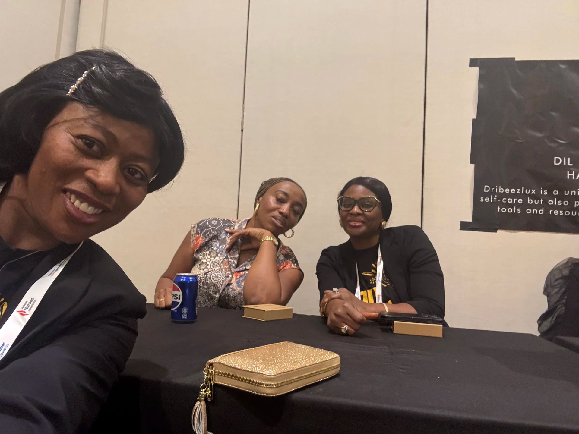 Three people seated at a table, smiling at a conference or event, with drinks and a small clutch in front.