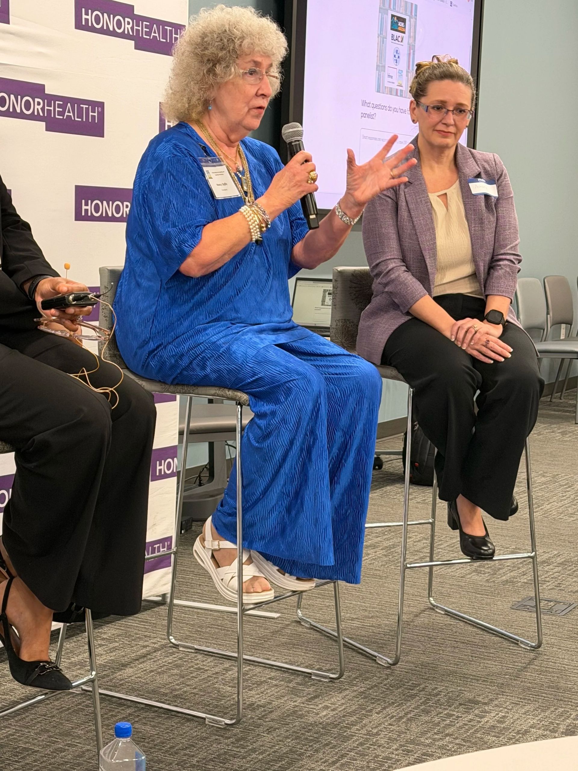 Two women seated on stools speaking at a panel, one in a bright blue dress gesturing with hands.