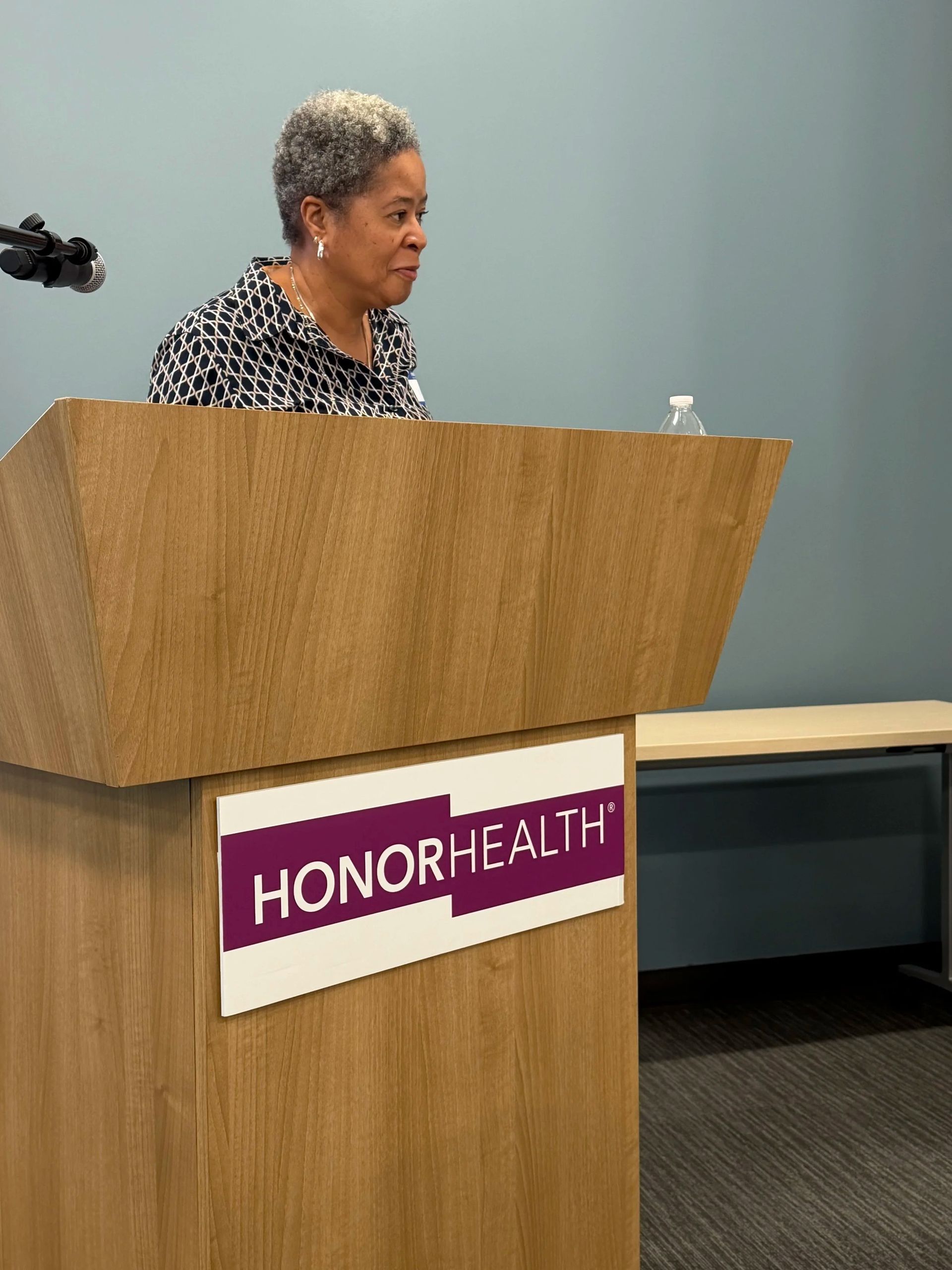 Woman speaking at a wooden podium with a HONOR HEALTH sign in a conference room.