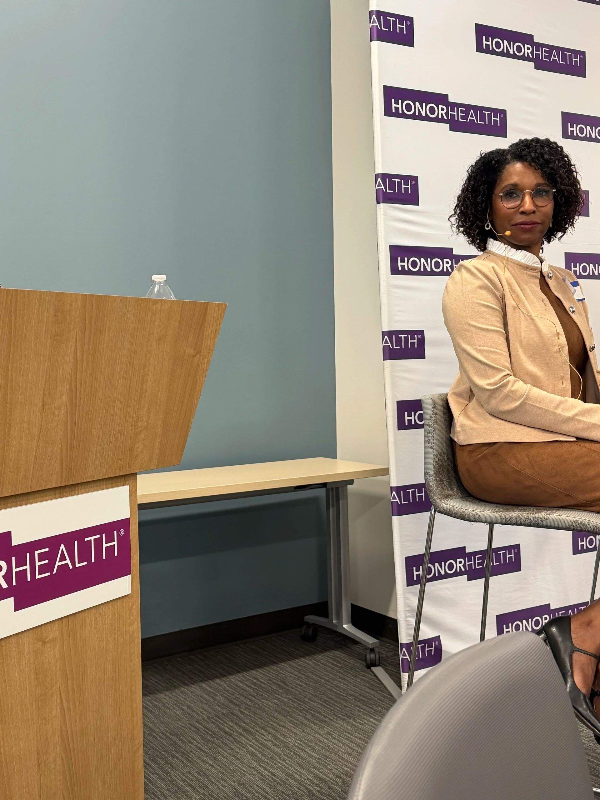 Woman seated beside a podium at a healthcare event, with a branded backdrop behind her.