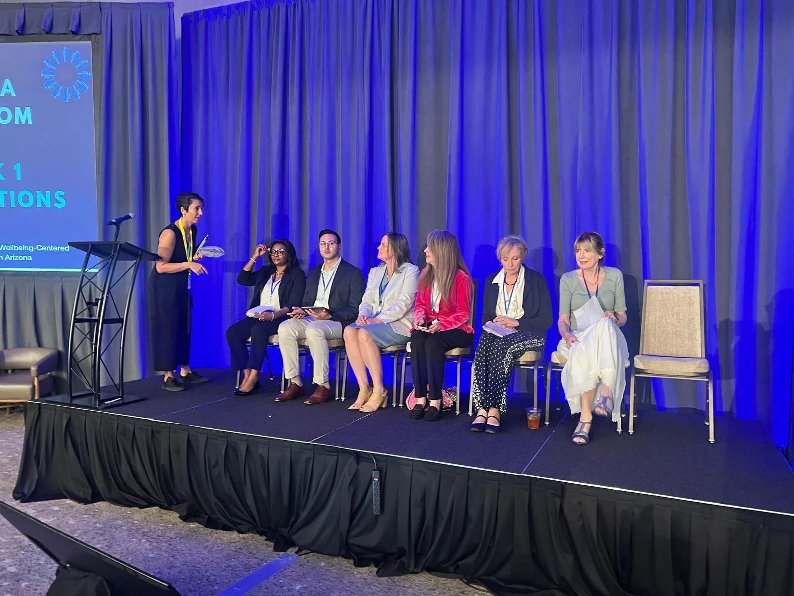 Panelists seated onstage beside a speaker, with blue curtains and a screen backdrop