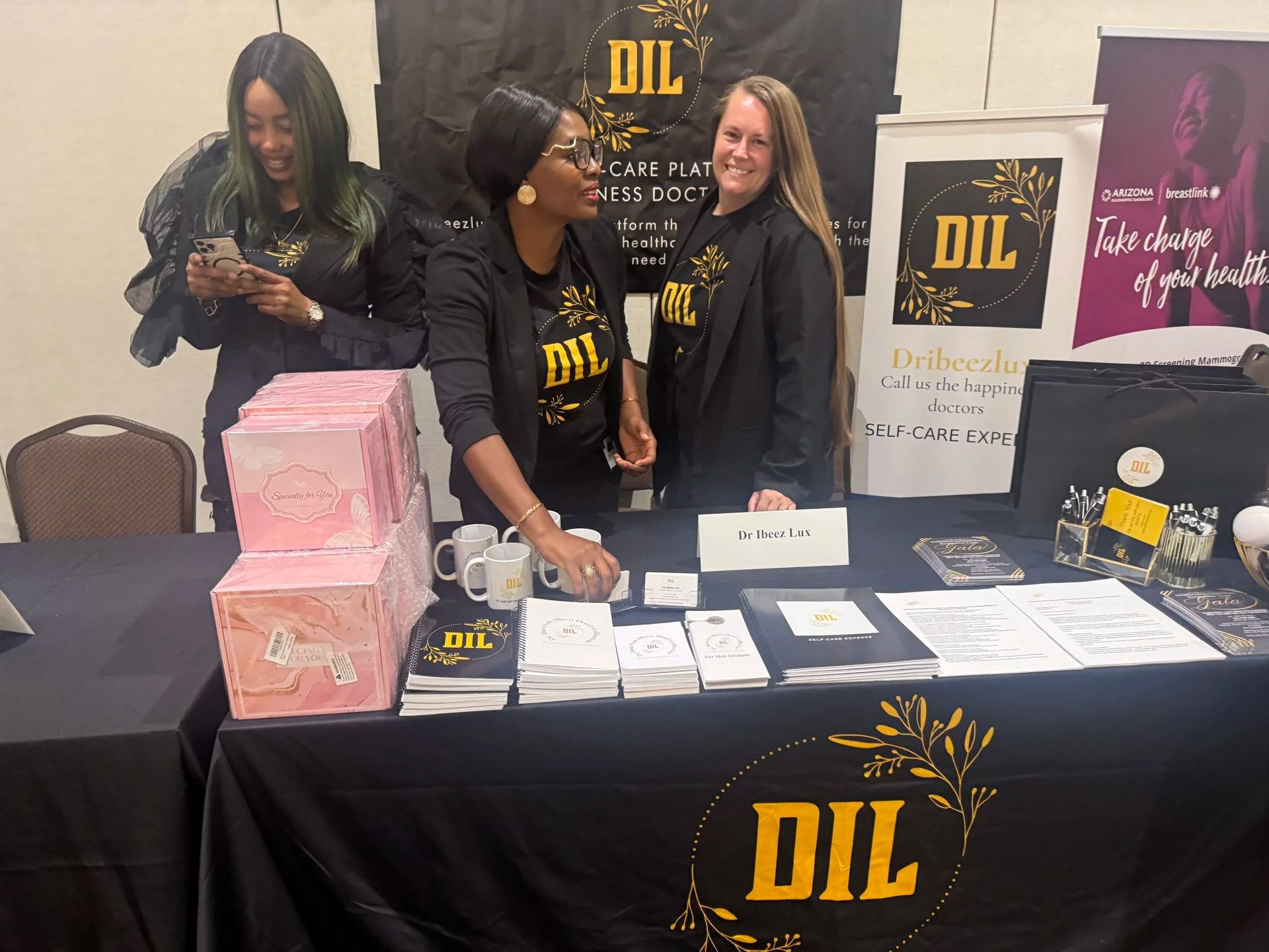 Three women at a DIL booth with brochures, books, and a banner at an indoor event