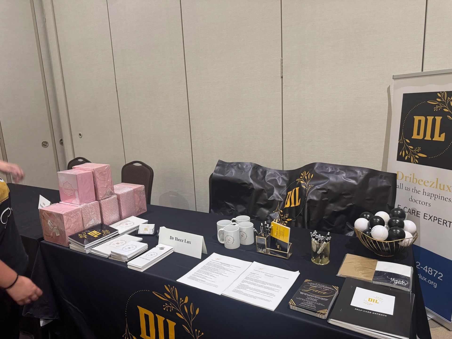 Black table display with pink gift boxes, black bags, small products, and a sign at an indoor event.