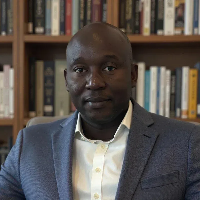 Man in a blue blazer seated in front of bookshelves, looking at the camera.