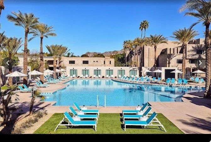 Resort pool with turquoise loungers, palm trees, and beige buildings under a bright blue sky