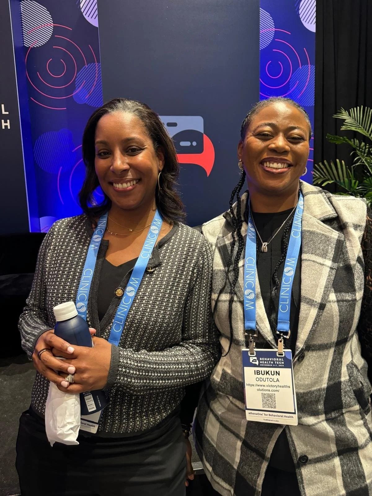 Two smiling people standing at a conference booth, wearing lanyards and holding microphones.