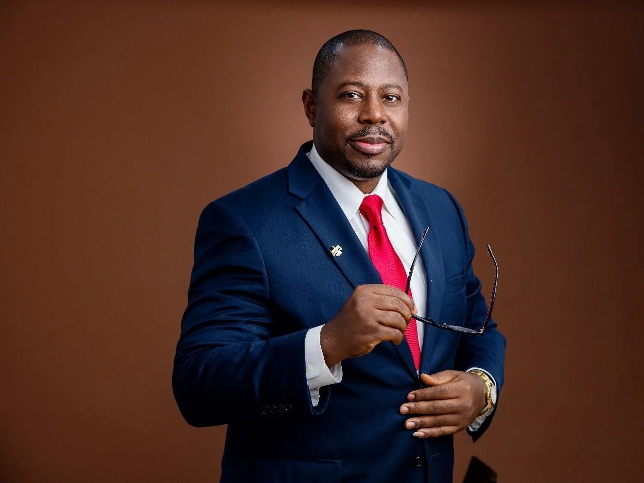 Man in navy suit and red tie holding glasses against a plain brown backdrop