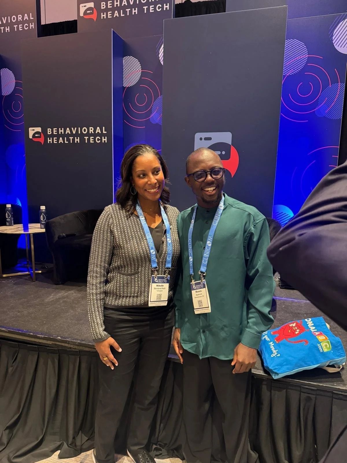 Two people smiling at a conference booth with Behavior Health Tech signage and a blue tote bag
