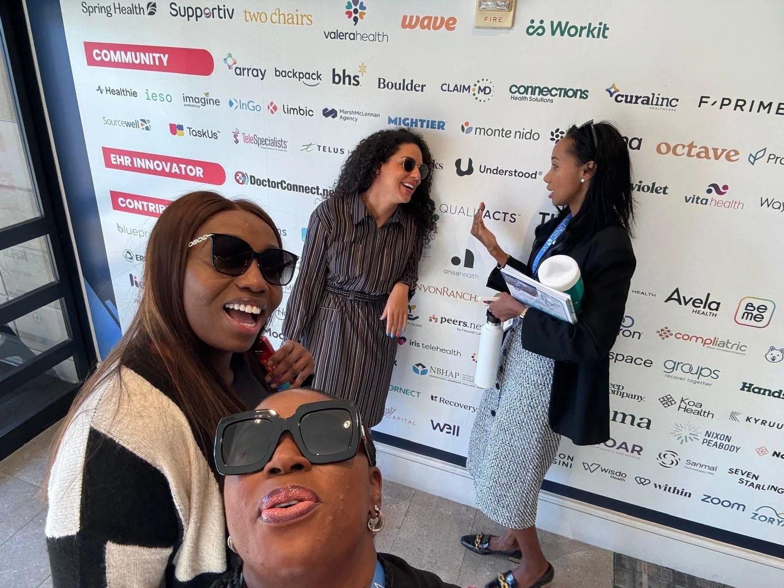 Three women posing in front of a branded event backdrop, smiling and talking in a bright room.