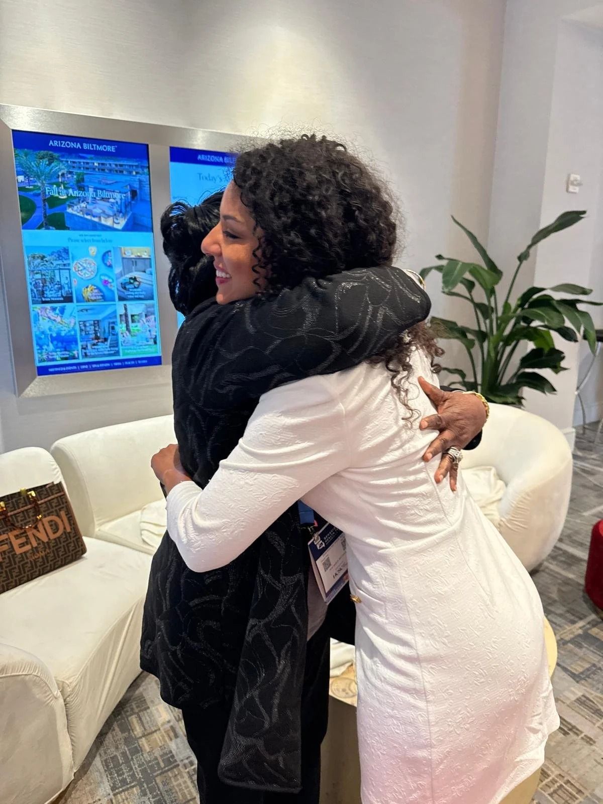 Two people hugging in a bright living room with white furniture and blue-lit screens.