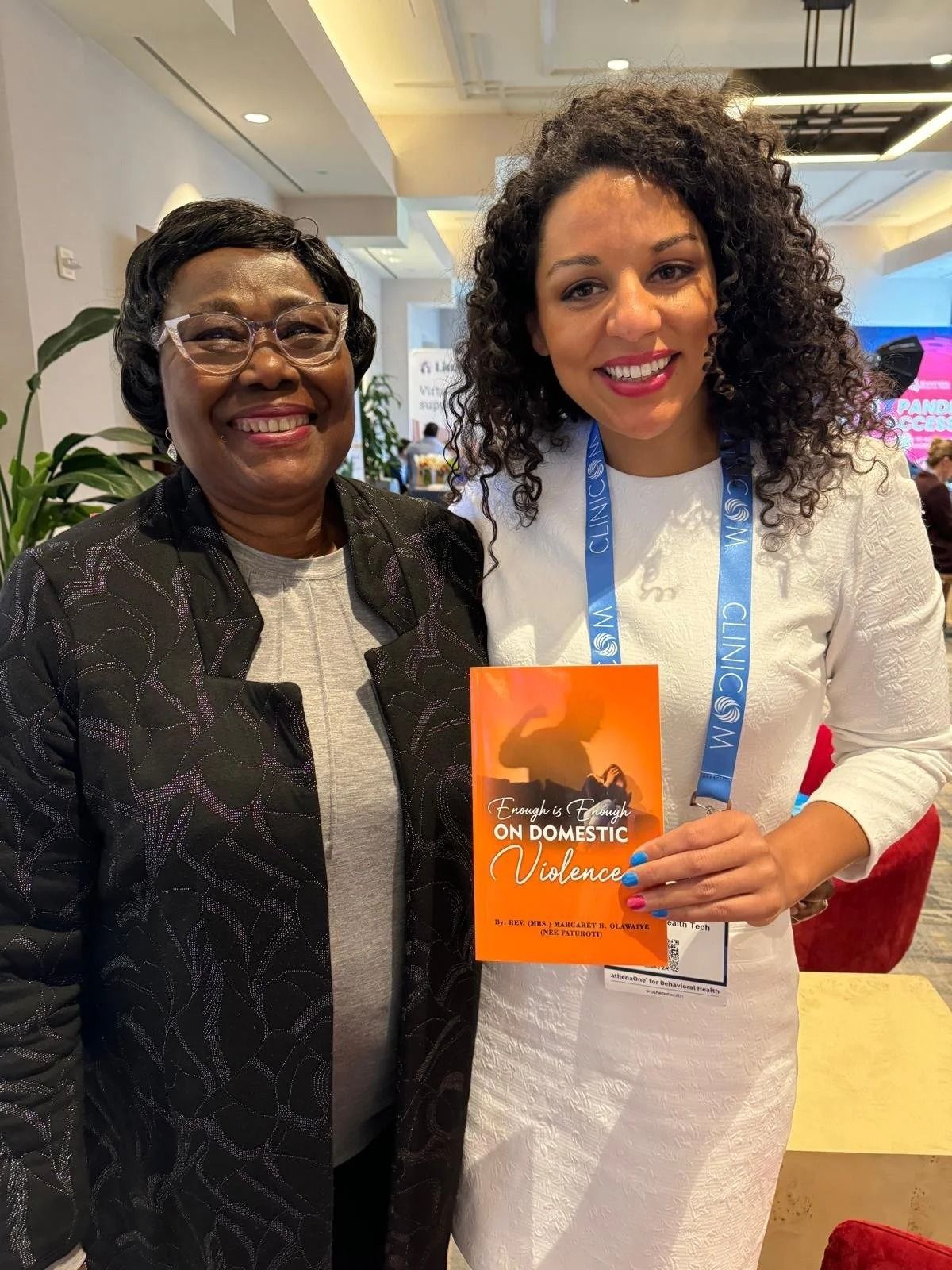 Two women smiling indoors, one holding an orange book with a blue lanyard around her neck.