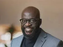 Smiling man in glasses and a gray blazer against a blurred indoor background