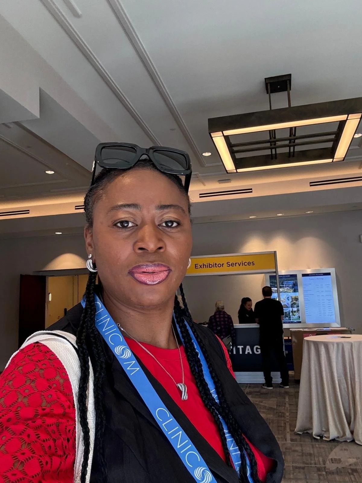 Woman with sunglasses on head and blue lanyard, standing indoors at a conference venue.