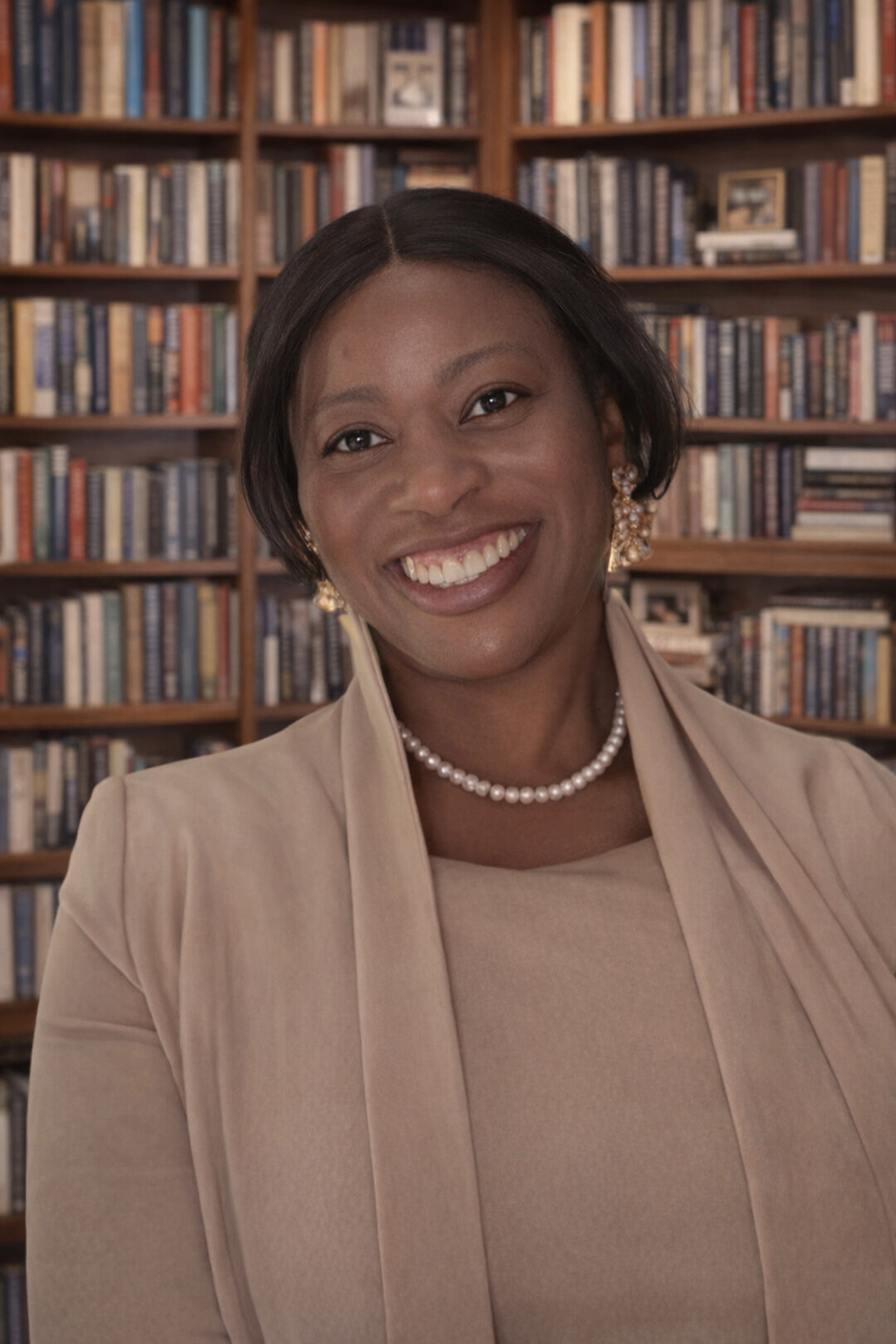 Smiling woman in beige outfit standing before a bookshelf backdrop
