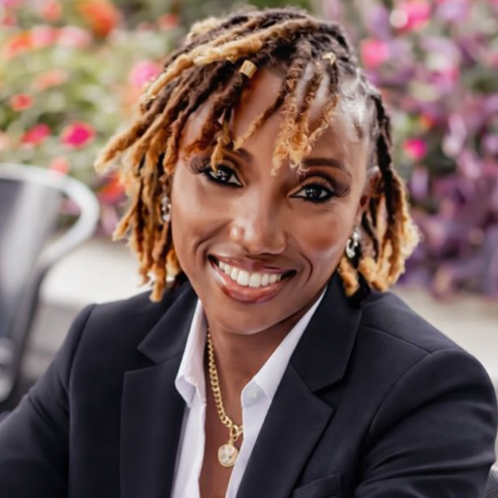 Smiling person in a dark blazer and white shirt, with blonde locs, seated against a blurred floral background