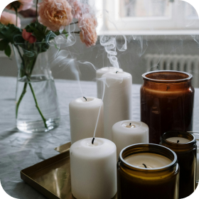 White candles and an amber jar candle on a tray beside flowers on a table, with smoke drifting upward.