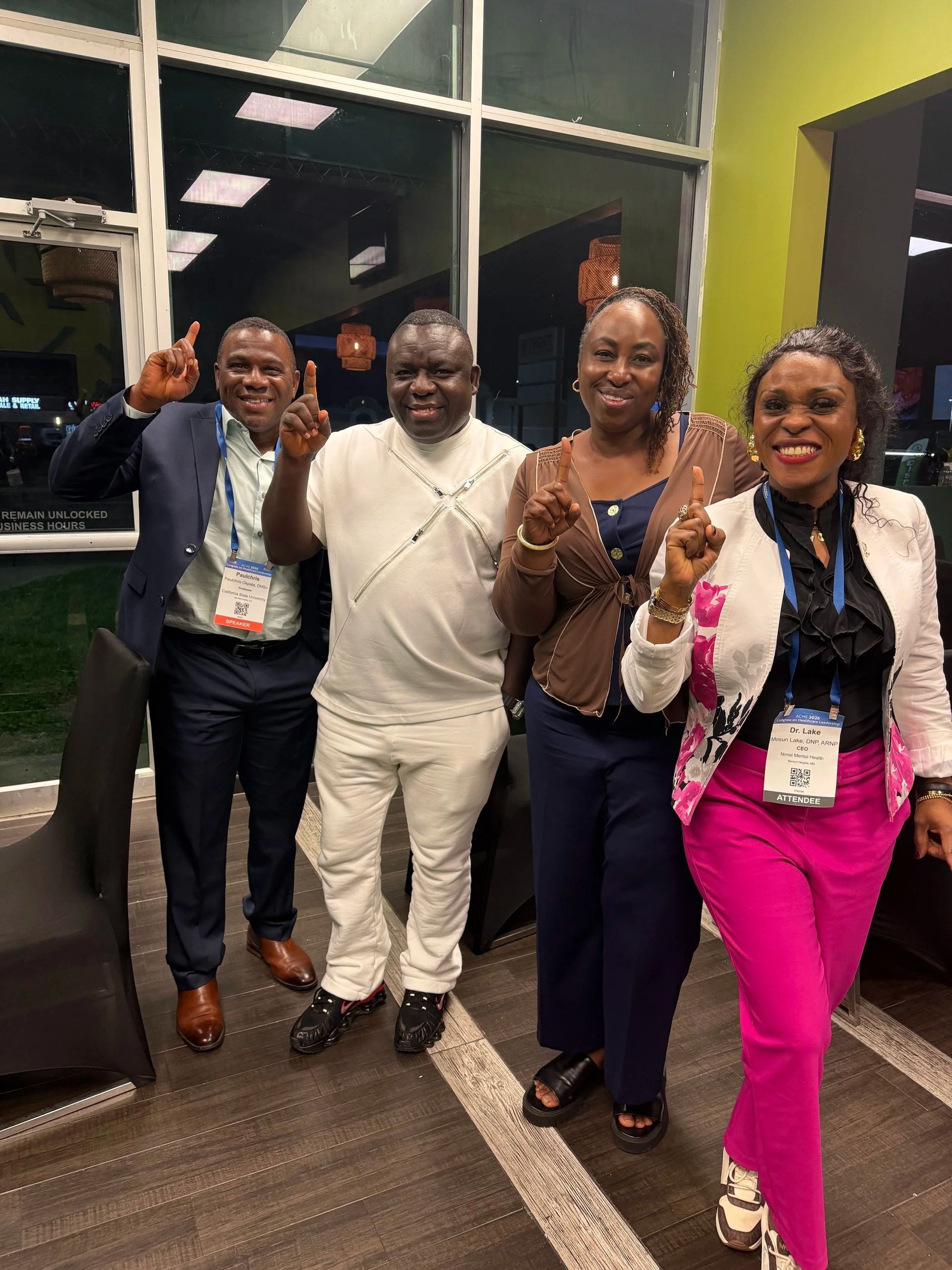 Four people smiling and holding up peace signs in a hallway, one in bright pink pants and black jacket.