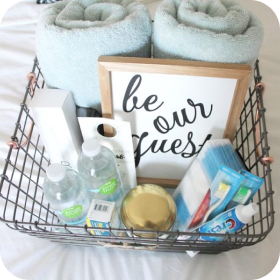Wire basket with toiletries, towels, and a “be our guest” sign for a guest room.