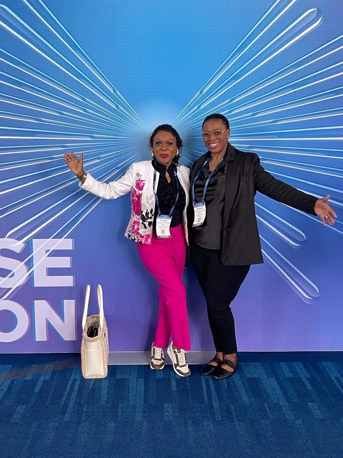 Two people posing with arms out in front of a blue event backdrop, one in pink pants and one in dark suit