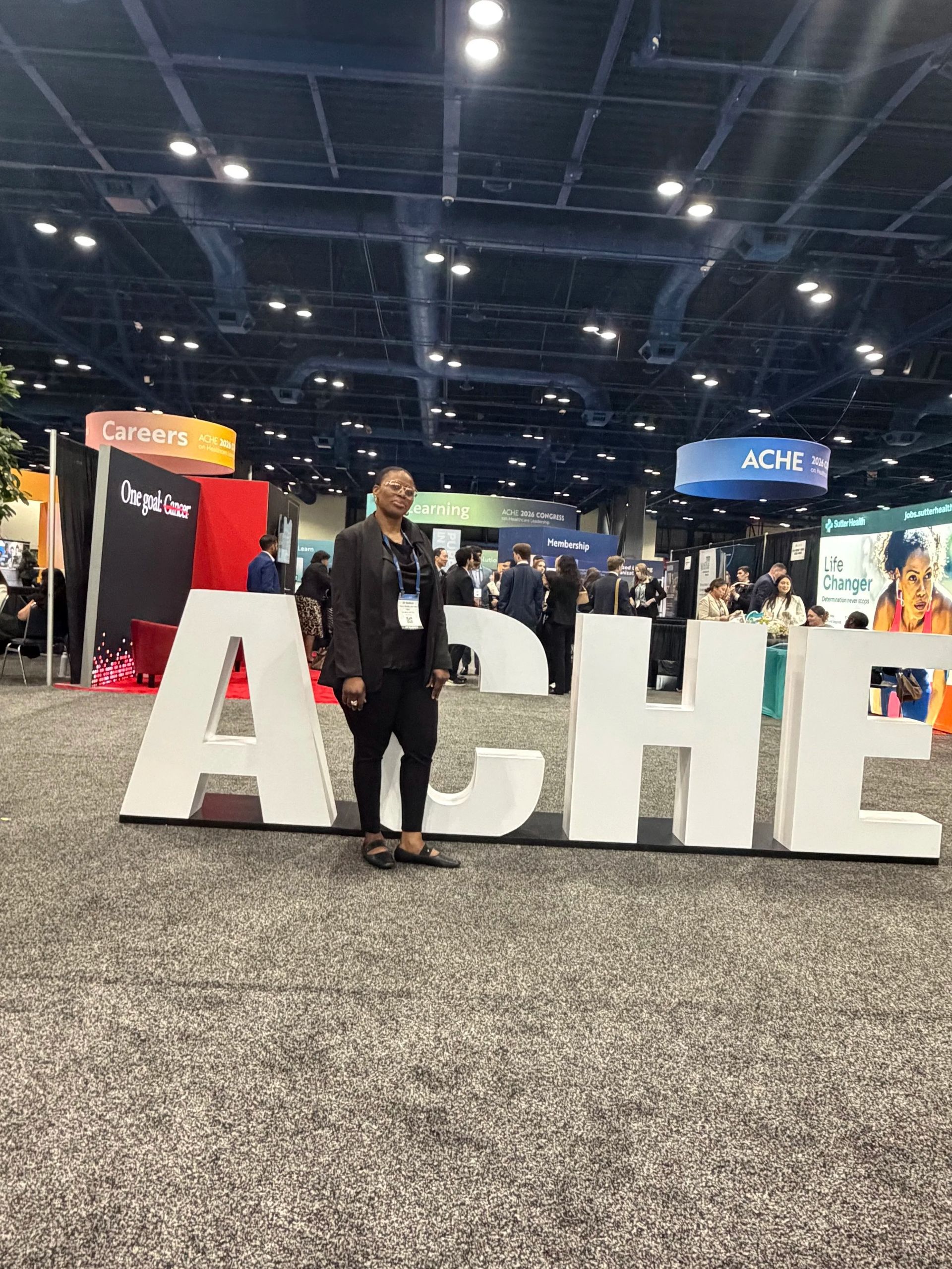 Person standing by large white “ACHE” letters at a busy indoor event venue.